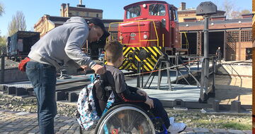 Junge im Rollstuhl und sein Einzelfallhelfer betrachten eine rote Lokomotive auf dem Rangierplatz im Museumshof